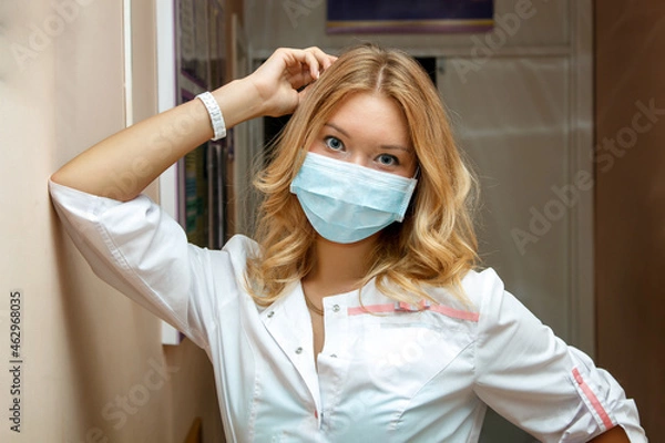 Obraz a young tired woman doctor in a mask and a white coat is leaning against the wall in the corridor of the hospital