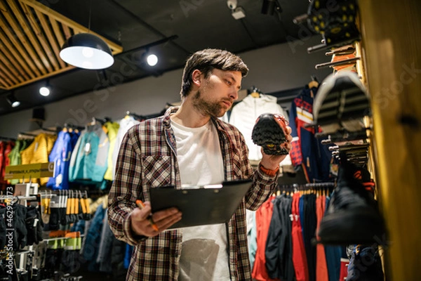 Fototapeta Sporting goods store owner with clipboard checking inventory. Tourist store manager working near showcase with hiking boots makes check list of orders. Salesman exhibit display of walking boots