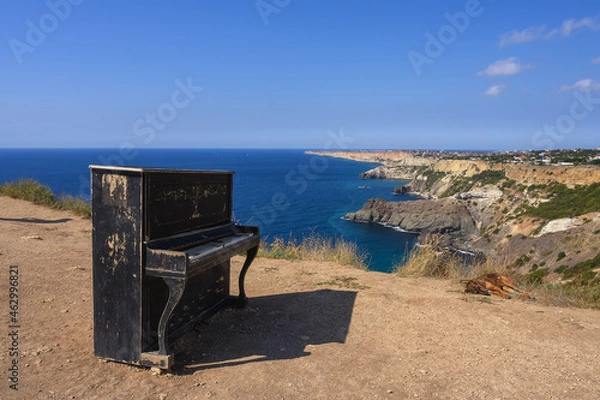 Fototapeta An old black piano and a dog lying next to him on a high rocky coast in the Crimea at Cape Fiolent. In the background there is a picturesque bay with blue water and blue sky. The city in the distance 