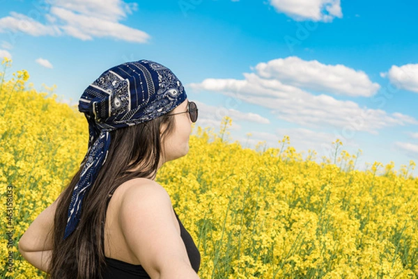 Fototapeta A girl in sunglasses and a bandana looks at the sky against the background of a blooming field