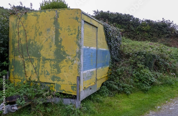Fototapeta Abandoned weathered colourful industrial trailer, overgrown with ivy left by a roadside. Towing hitch Flaking yellow, blue and white paintwork. Landscape image with space for text. UK.