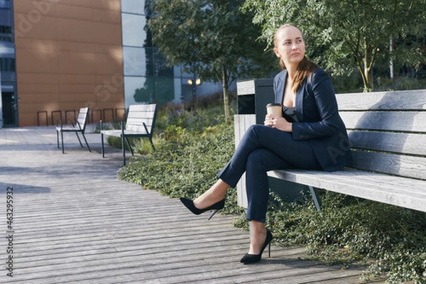 Fototapeta Pensive european business woman has coffee break, sits outside of office on bench with takeaway cup in hand, looks aside, wears business suit, high heels and deep neckline blouse, has ponytail.