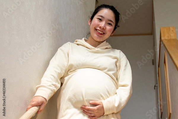 Obraz A young pregnant Asian woman with smile goes down the stairs carefully by using handrails in a house