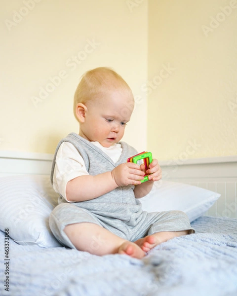 Fototapeta Funny infant baby boy holding magnetic constructor toy and looking away in bedroom. Adorable little child in cute outfit enjoying playtime, sitting on bed at home or kindergarten