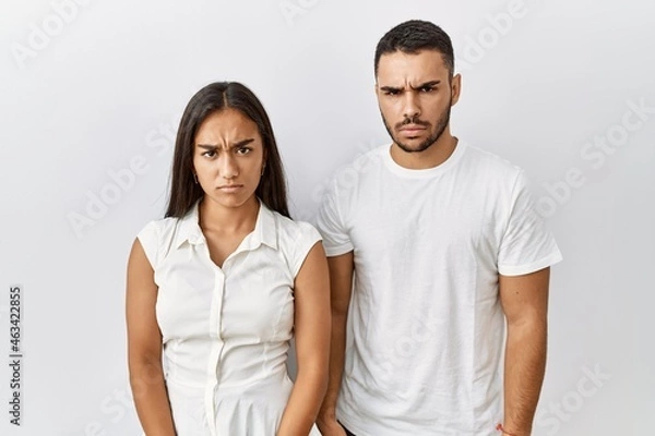 Fototapeta Young interracial couple standing together in love over isolated background skeptic and nervous, frowning upset because of problem. negative person.