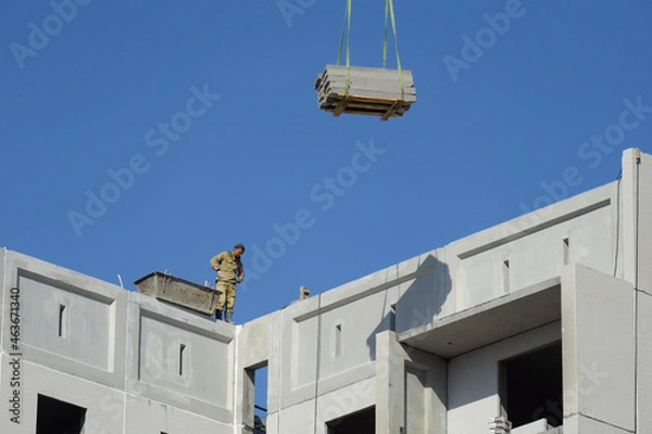 Fototapeta Construction of a multi-storey building. An unsecured builder stands on the edge and looks down. The plates are lifted up by a crane.