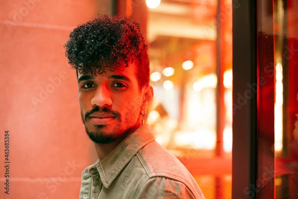 Fototapeta Portrait of young biracial curly haired man with orange neon light reflection on face. Looking towards the camera. Millennial with beard and earring. Horizontal image.