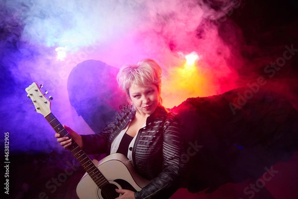 Fototapeta Self-assured adult blonde middle age woman with black angel wings and guitar like a rock musician. Model posing in studio on black background. Dangerous cupid on Valentine's day