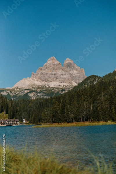 Obraz Auf dem Bild ist ein hoher Berg vor einem blauen Himmel zu sehen. Am Fuße des Berges liegt ein ruhiger See, umgeben von einem dichten Wald.
