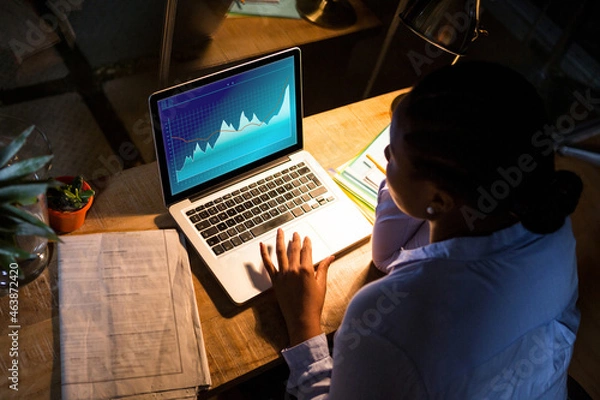 Fototapeta Biracial businesswoman sitting at desk using laptop with statistical data on screen