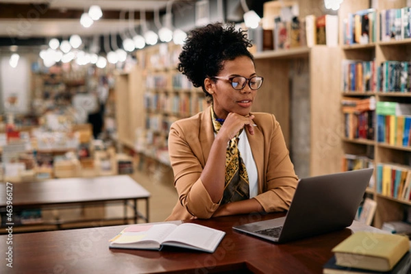 Fototapeta African American female professor using laptop while doing a research at university library.