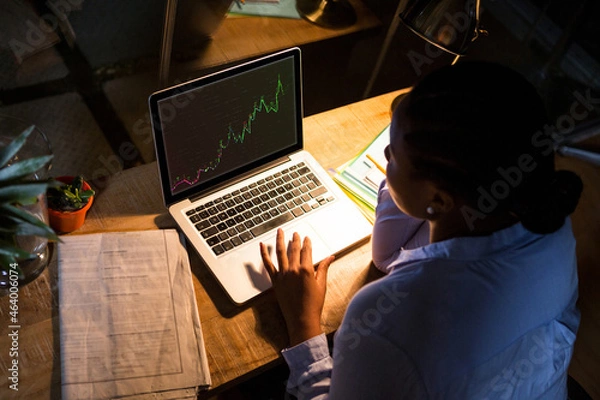 Fototapeta Biracial businesswoman sitting at desk, using laptop with statistical data on screen