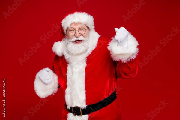 Fototapeta Photo of impressed lucky man pensioner dressed santa claus costume rising fists smiling isolated red color background