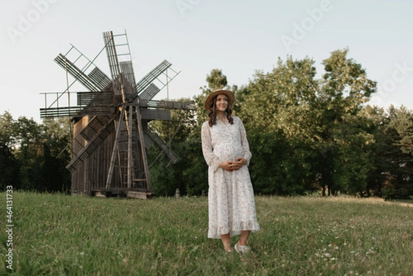 Fototapeta A full-length portrait of a pregnant woman in the windmill background who is holding her belly with her hands on the farm. A happy pregnancy. A lady in a dress and a hat at the meadow in the evening.
