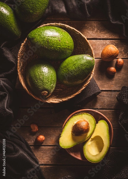 Fototapeta Some open and close ripe avocados in wooden bowls on a dark brown wooden table with black patterned and avocado seeds around. Dark food.