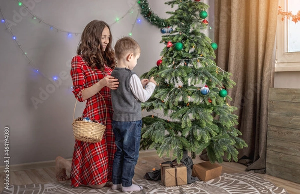 Obraz Mom with her child is decorating together the Christmas tree with toys and balls. Concept of family preparation for the New Year, cozy atmosphere and festive mood