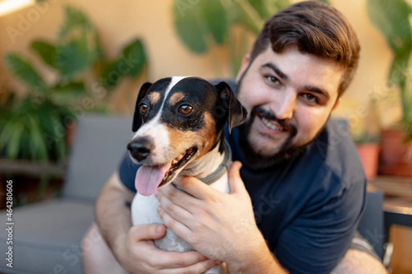 Fototapeta Friendly young man hugging his cute grocer dog with both hands and looking at camera. The dog has the tongue out and is looking away