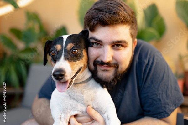 Fototapeta Friendly young man hugging his cute grocer dog with both hands and looking at camera. The dog has the tongue out and is looking away