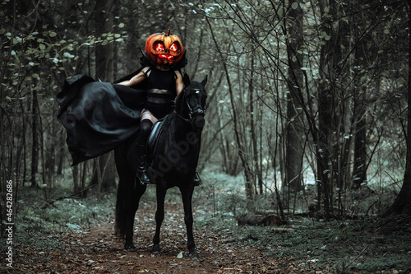 Fototapeta the rider is sitting astride a horse. a girl in a long black raincoat with a pumpkin mask with burning eyes. Creepy cosplay shooting for Halloween in the autumn forest. a cloak fluttering in the wind
