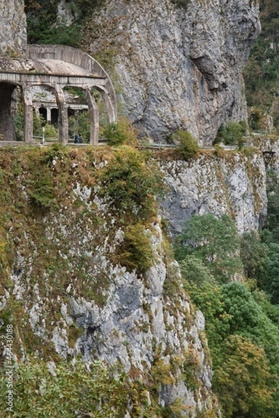 Fototapeta The mountain road goes into a gorge between a sheer cliff and a cliff. The river is deep at the bottom of the gorge.