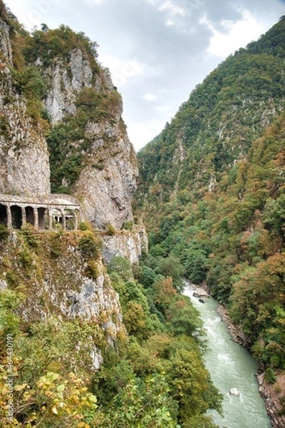 Fototapeta The mountain road goes into a gorge between a sheer cliff and a cliff. The river is deep at the bottom of the gorge.