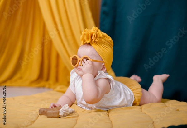 Obraz A fashionable baby girl is resting in yellow glasses. Cute girl posing in the studio in a yellow beanie cap