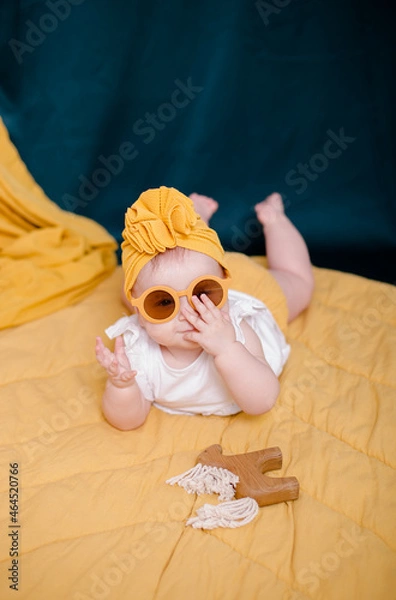 Obraz A fashionable baby girl is resting in yellow glasses. Cute girl posing in the studio in a yellow beanie cap