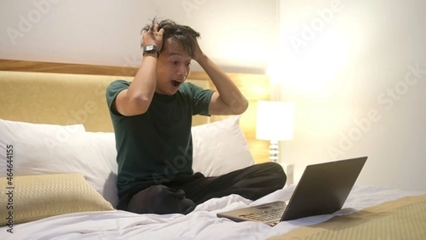 Fototapeta Angry Asian man is working with his laptop on his bed. Concept of freelancer problems.