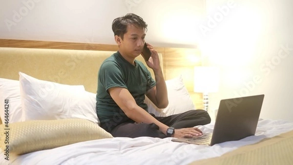 Obraz Happy Asian man working with laptop while holding smartphone on bed