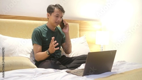 Obraz Happy Asian man working with laptop while holding smartphone on bed