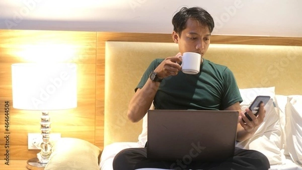Fototapeta Happy creative Asian man working with laptop while holding cup on mattress