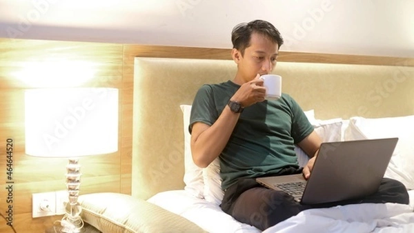 Fototapeta Happy creative Asian man working with laptop while holding cup on mattress