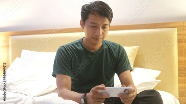 Fototapeta Sleepy Asian man is using smartphone while lying on the bed.