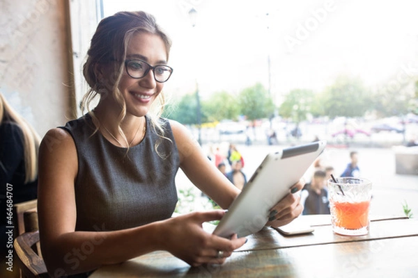 Obraz Smiling caucasian woman in glasses reading newsletter on digital gadget while sitting in coffee shop. Cheerful female student having online learning via touch pad