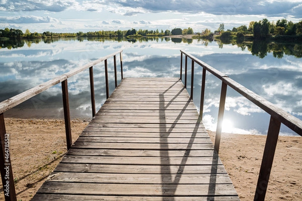 Fototapeta Panorama of the pier by a quiet lake on a sunny day. A wooden pier at the water surface, clouds are reflected in the water, and a forest grows around.