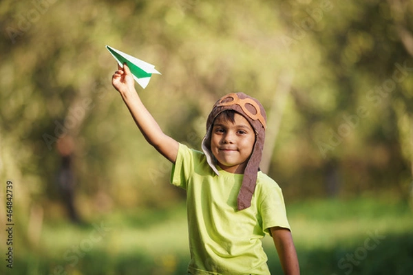 Fototapeta cute little Latin American boy in a pilot's hat plays as a pilot, launching a paper airplane into air. child is happy about the flight of the airplane. Children's active outdoor game. Dynamic image