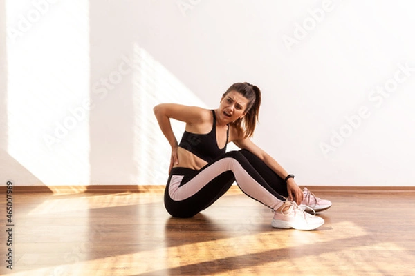 Fototapeta Athletic female frowning from pain in lower back, muscle strains and ligament sprains problem, wearing black sports top and tights. Full length studio shot illuminated by sunlight from window.