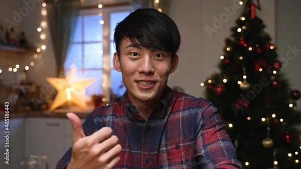 Obraz Close up portrait of Asian korean happy man looking camera speaking on video call with friend and having good talk sending greetings sitting near Christmas tree in room. New Year concept