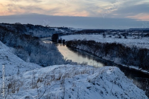 Fototapeta A beautiful winter view from the hill to the river with frosty trees growing along the banks. Winter landscape at sunset