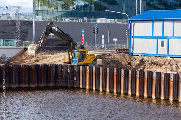 Fototapeta Excavator on a construction site with a foundation pit reinforced with a steel sheet pile. Modern construction of road junction and buildings