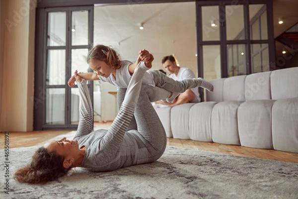 Fototapeta Jolly mother is doing gymnastics with girl on carpet while father and son are using notebook