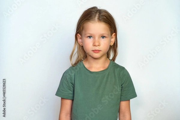 Obraz serious child looks at the camera. place for text. Portrait of a girl on a white background. Collected hair and green T-shirt. Cute smile happy smile. A cheerful preschool 
