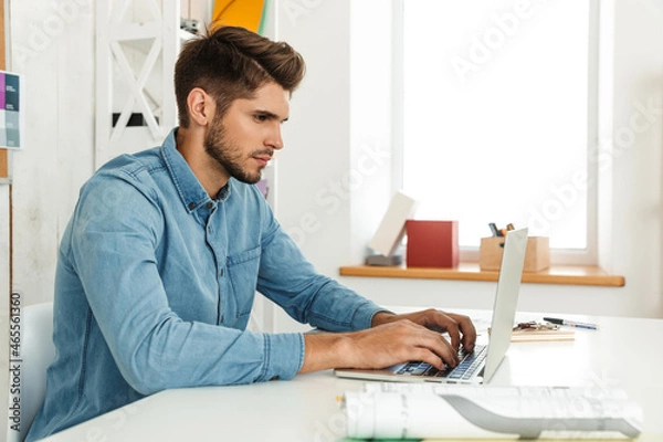 Fototapeta Young white man working with laptop while sitting at desk in office