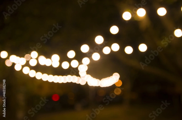 Fototapeta Blurred abstract background, backyard illumination, light in the evening garden, electric Defocused garland. Lamp garland of light bulbs on trees illuminate night scene