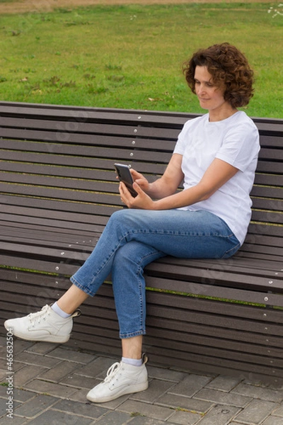 Fototapeta A 45-50-year-old woman is sitting on a bench with a mobile phone in her hands