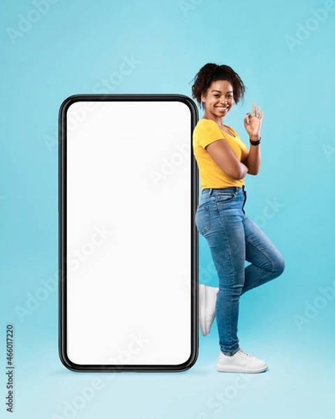 Fototapeta Cheerful black woman standing near big cellphone with empty white screen, gesturing okay on blue background, mockup