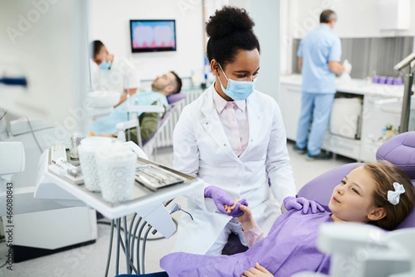 Fototapeta African American dentist holds little girl's hand and consoles her during dental procedure at dentist's office.