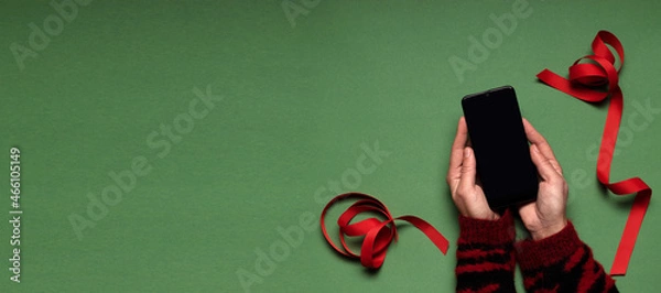 Fototapeta Top view.Woman wearing red sweater and holding smart phone with blank screen, red xmas ribbon on the green surface.Online buying presents,gifts
