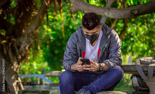 Fototapeta Man with a mask sitting on a bench with his phone, young man with a mask sitting with a cell phone, near a young man with a mask texting