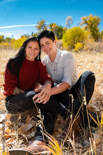 Fototapeta Lesbian couple holding each other in a field during autumn surrounded by leaves and smiling. 